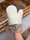 A cream-colored silicone oven mitt with a textured non-stick surface and a tan wrist strap featuring a circular embroidered logo that reads "Dipped by Ryder", shown being worn on a person's hand over a rustic wooden table background.
